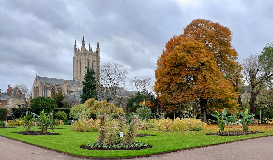A cultivated garden area featuring a variety of plants and vegetables, including leafy greens, corn, and flowering shrubs, arranged in raised beds and borders. The garden has a weathered pathway made 